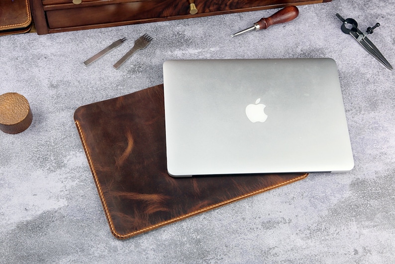 May include: A brown leather laptop sleeve with a laptop inside. The laptop is silver and has an Apple logo on the back.