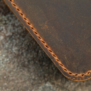 May include: A dark brown leather object with a smooth, matte surface and visible scuff marks. The edge is rounded and secured with a thick, light brown waxed thread stitched in a consistent, repeating pattern along the perimeter of the material.