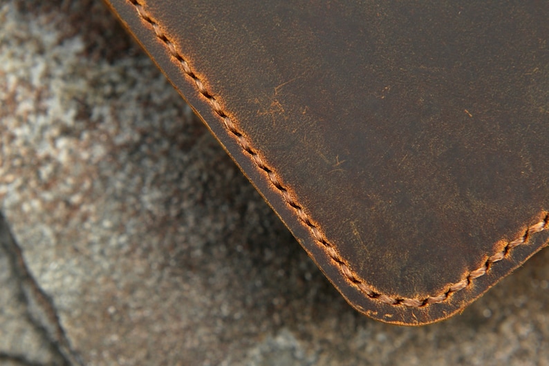 May include: A dark brown leather object with a smooth, matte surface and visible scuff marks. The edge is rounded and secured with a thick, light brown waxed thread stitched in a consistent, repeating pattern along the perimeter of the material.