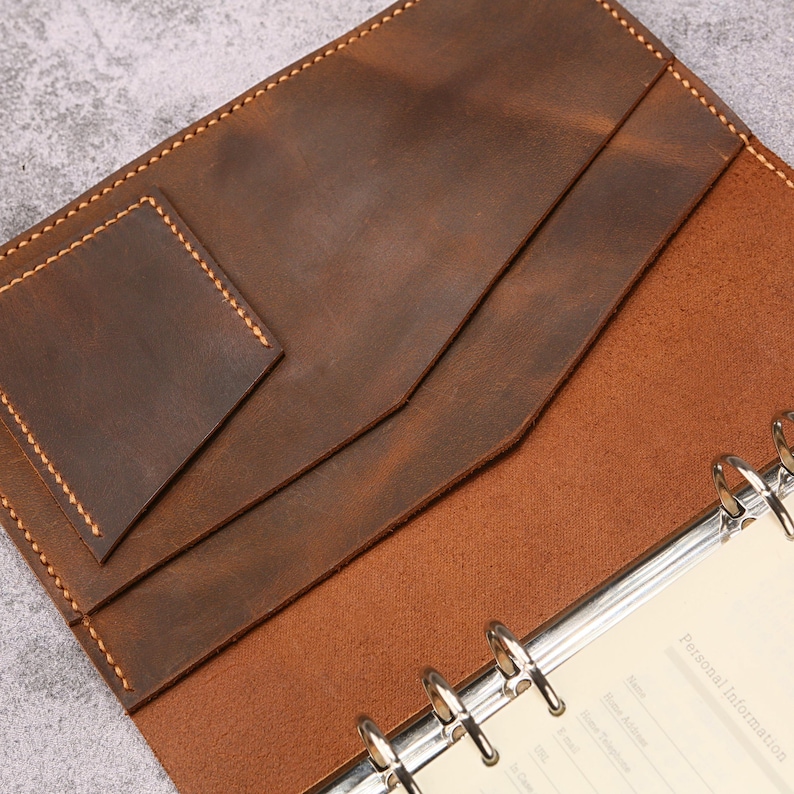 May include: A brown leather planner cover featuring a six-ring metal binder mechanism and interior pockets. The leather has visible stitching along the edges and a smooth, matte finish. A page with the printed text Personal Information is visible.