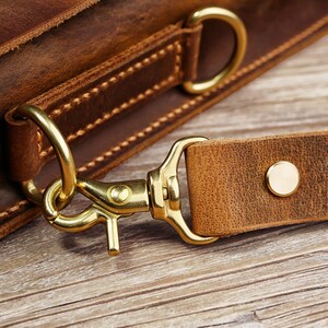 May include: Close-up of a brown leather bag with a brass hook and strap. The strap is attached to the bag with a brass ring.