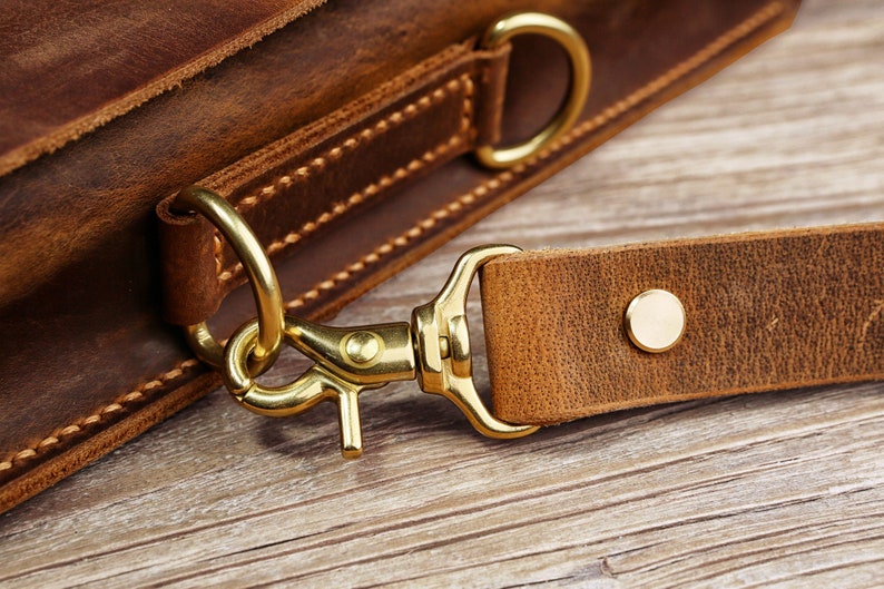 May include: Close-up of a brown leather bag with a brass hook and strap. The strap is attached to the bag with a brass ring.