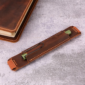 May include: A brown leather pen holder with a black clip and a green pen inside. The pen holder is stitched with light brown thread and has a brown elastic band. A brown leather notebook is in the background.
