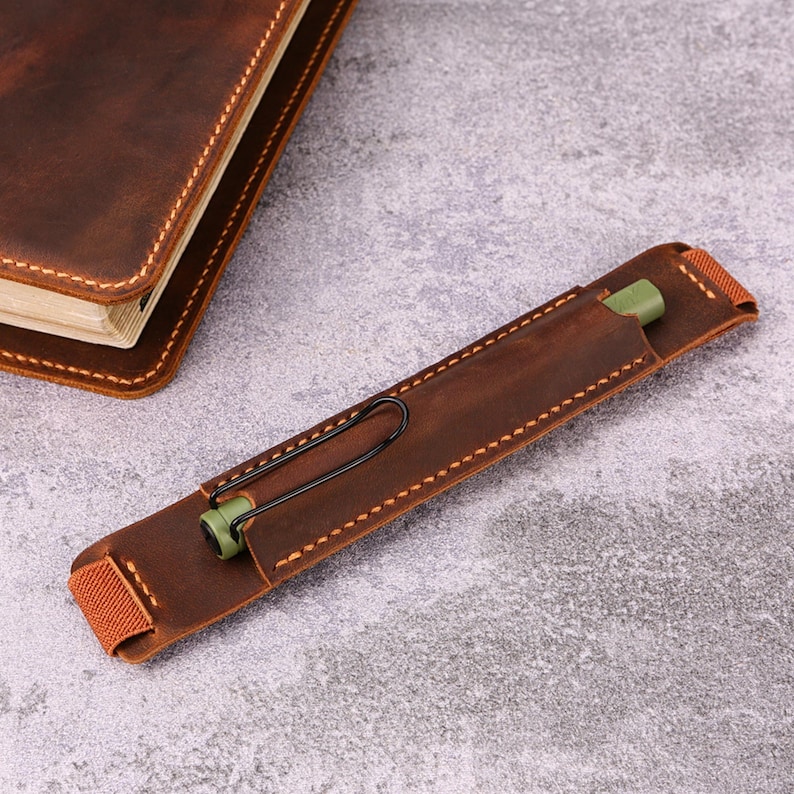 May include: A brown leather pen holder with a black clip and a green pen inside. The pen holder is stitched with light brown thread and has a brown elastic band. A brown leather notebook is in the background.