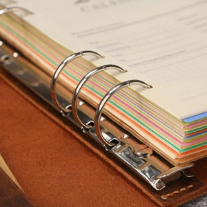 May include: A brown leather binder featuring a silver metal six-ring mechanism. The interior contains cream-colored pages with thin, horizontal, multi-colored stripes along the edges, including shades of green, orange, blue, and yellow.