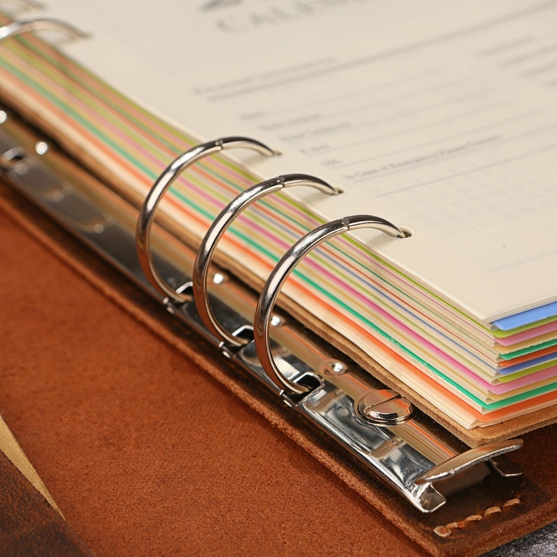 May include: A brown leather binder featuring a silver metal six-ring mechanism. The interior contains cream-colored pages with thin, horizontal, multi-colored stripes along the edges, including shades of green, orange, blue, and yellow.