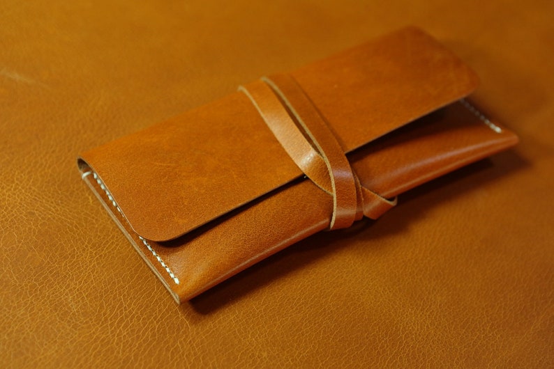 May include: A brown leather wallet with a strap closure. The wallet is folded in half and the strap is tied around it.