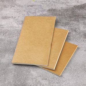 Könnte beinhalten: Drei braune Notizbücher, die übereinander gestapelt sind. Die Notizbücher haben einen strukturierten Papierumschlag und weiße Kanten.