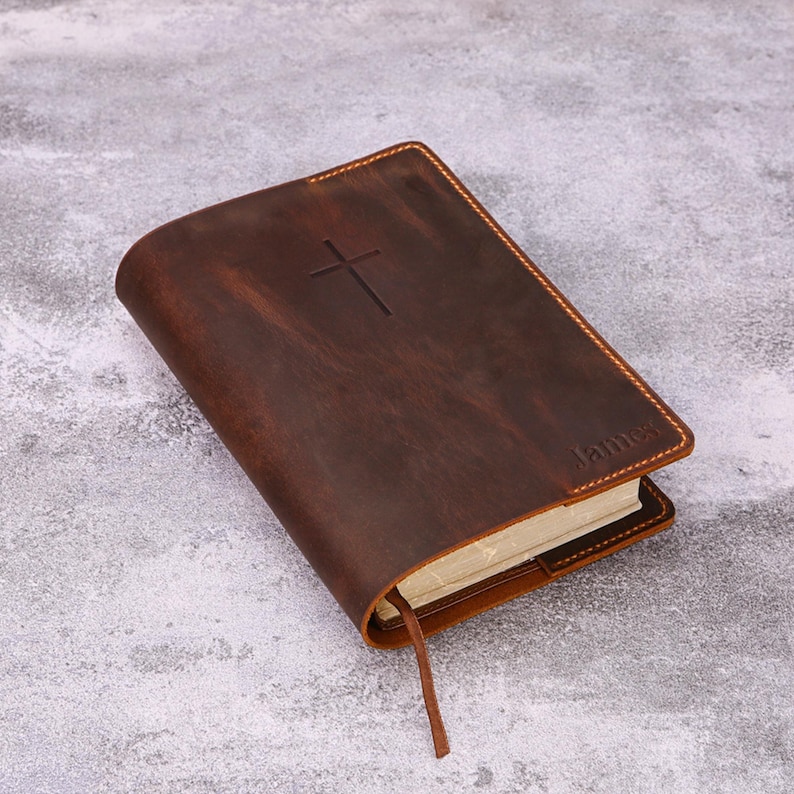 May include: A brown leather-bound book with a cross embossed on the cover. The word "James" is visible on the bottom right corner. The book has a sewn edge and a brown ribbon bookmark.