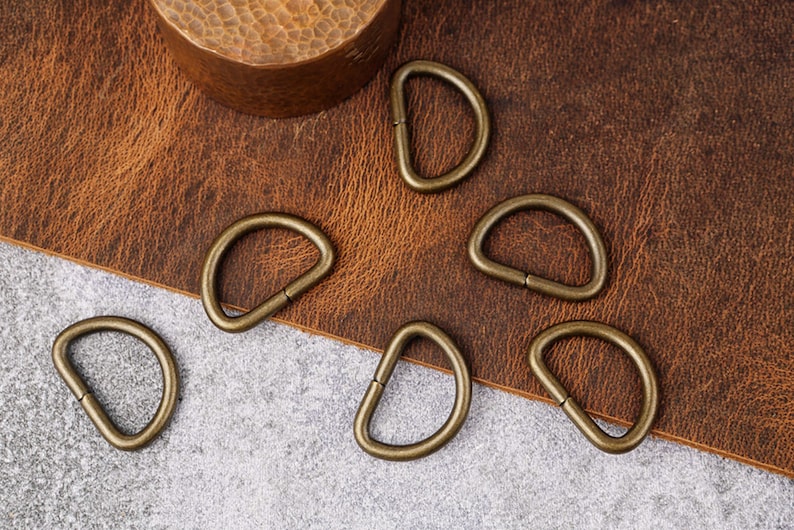 May include: Six antique gold-toned metal D-rings on a brown leather surface and a gray surface. The D-rings are approximately 1 inch in diameter.