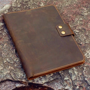 May include: A brown leather folder with a flap closure and two brass poppers. The folder has a distressed look and is lying on a rough, textured surface.