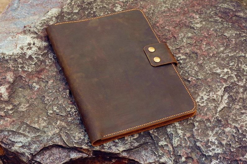 May include: A brown leather folder with a flap closure and two brass poppers. The folder has a distressed look and is lying on a rough, textured surface.