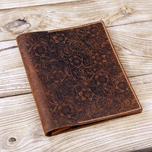 May include: A brown leather journal with a floral embossed design. The journal is closed and lying on a wooden surface.