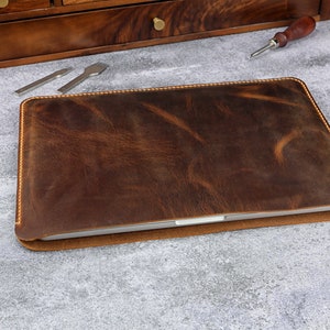 May include: A brown leather laptop sleeve with a stitched edge. The sleeve is lying on a gray surface.