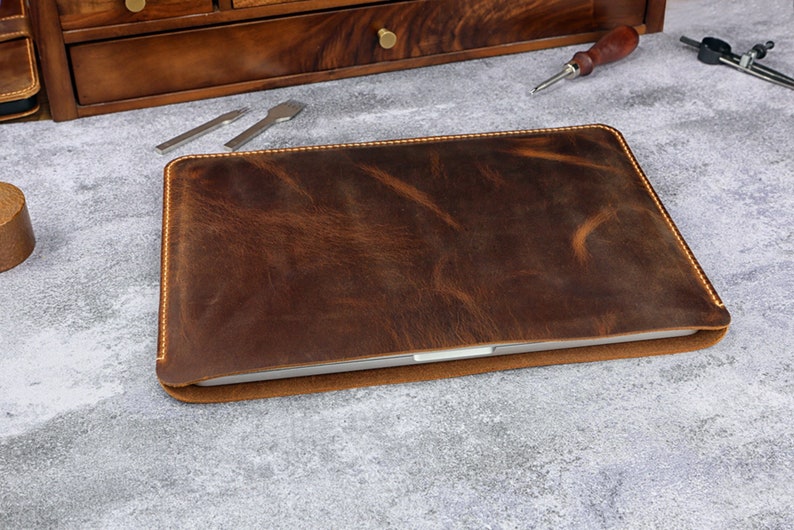 May include: A brown leather laptop sleeve with a stitched edge. The sleeve is lying on a gray surface.