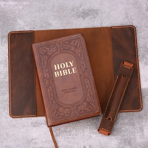 May include: A brown leather Bible with the words "Holy Bible" and "King James Version" embossed on the cover. The Bible is open, revealing a bookmark. It rests on a larger leather cover with a matching pen holder.