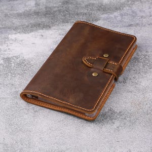 May include: A brown leather journal with a strap closure. The journal is rectangular and features a stitched border. The leather has a distressed appearance, with variations in color. The closure is secured with a leather strap and two brass-colored rivets.