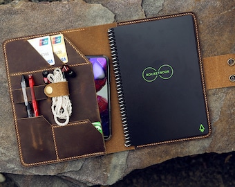 Könnte beinhalten: Eine braune Leder-Notizbuchhülle mit mehreren Taschen und einem integrierten Gummiband. Die Hülle trägt das Rocketbook-Logo auf der Vorderseite und enthält ein schwarzes Notizbuch mit Spiralbindung.
