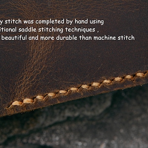 Op de afbeelding: Close-up van bruin leer met handgestikte details. De beige draad cre&euml;ert een contrasterend patroon. Tekst luidt: "Elke steek is met de hand voltooid met behulp van traditionele zadelstitchingstechnieken." Het leer heeft een verweerde uitstraling.