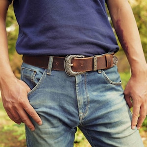 Pode incluir: Um cinto de couro castanho com uma fivela prateada decorativa. O cinto é usado com jeans azuis e uma camisa azul marinho. A fivela tem um design ornamentado. O cinto tem aproximadamente 3,8 cm de largura.