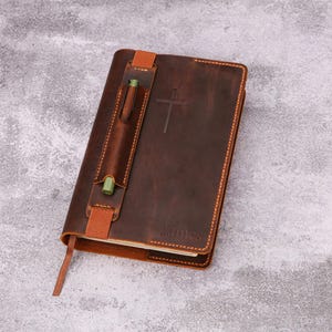 May include: A brown leather journal with a pen holder and a cross embossed on the cover. The journal has a bookmark and the name "James" is embossed on the lower right corner. The pen holder is secured with a brown strap.