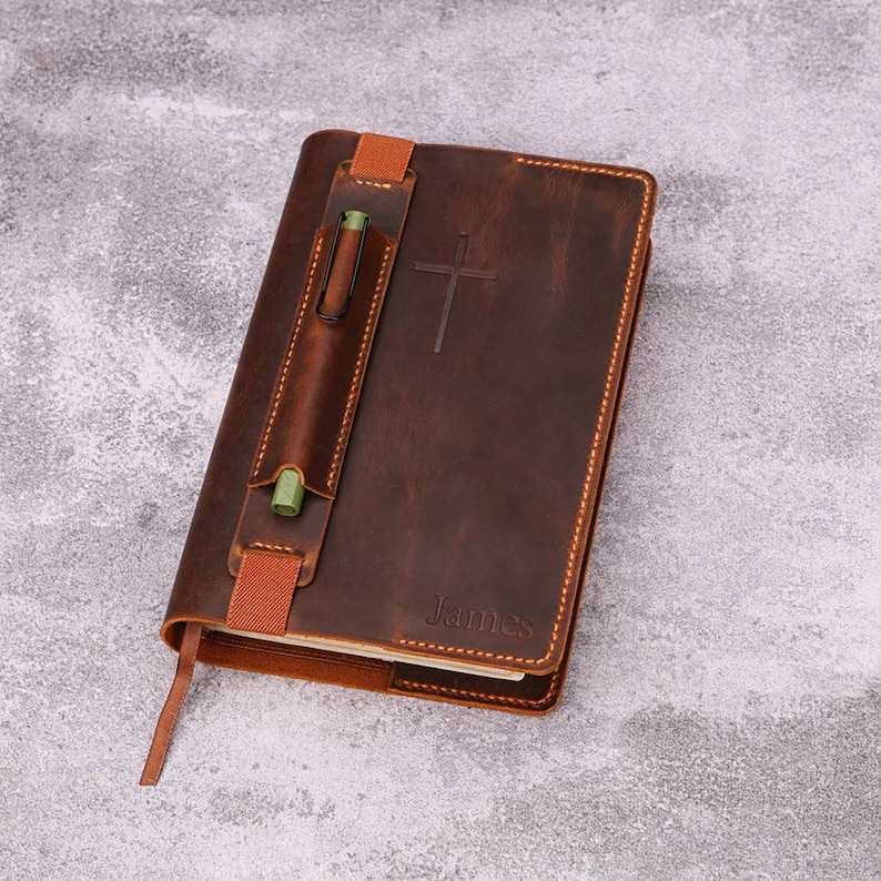 May include: A brown leather journal with a pen holder and a cross embossed on the cover. The journal has a bookmark and the name "James" is embossed on the lower right corner. The pen holder is secured with a brown strap.
