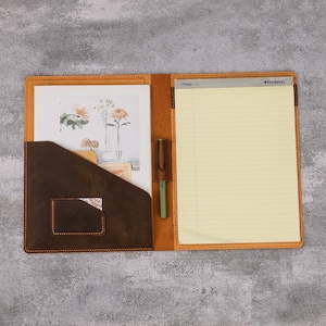 May include: Open leather portfolio in brown with a lighter tan interior. The left side has a dark brown pocket with a small card slot. A notepad and pen are visible inside. A floral print is in the left pocket.