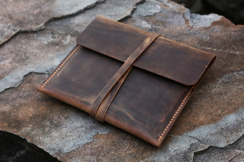 Puede incluir: Una funda de cuero marr&oacute;n de estilo sobre con cierre de correa. La funda tiene un aspecto desgastado y est&aacute; cosida con hilo marr&oacute;n claro.