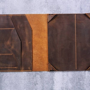 May include: A dark brown leather portfolio case featuring a smooth exterior finish with visible orange stitching. The interior includes a left-side panel with multiple pockets and a right-side panel with corner tabs for securing a tablet or notebook.
