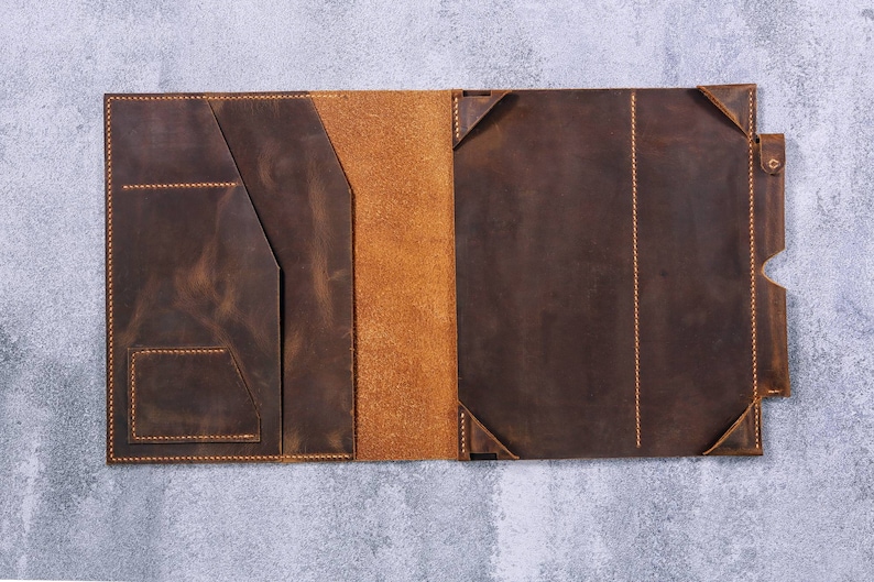 May include: A dark brown leather portfolio case featuring a smooth exterior finish with visible orange stitching. The interior includes a left-side panel with multiple pockets and a right-side panel with corner tabs for securing a tablet or notebook.