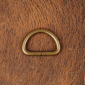 May include: A gold-toned metal D-ring, also known as a split ring, is shown against a brown leather background.