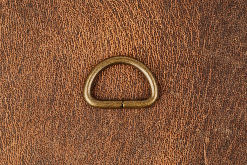 May include: A gold-toned metal D-ring, also known as a split ring, is shown against a brown leather background.