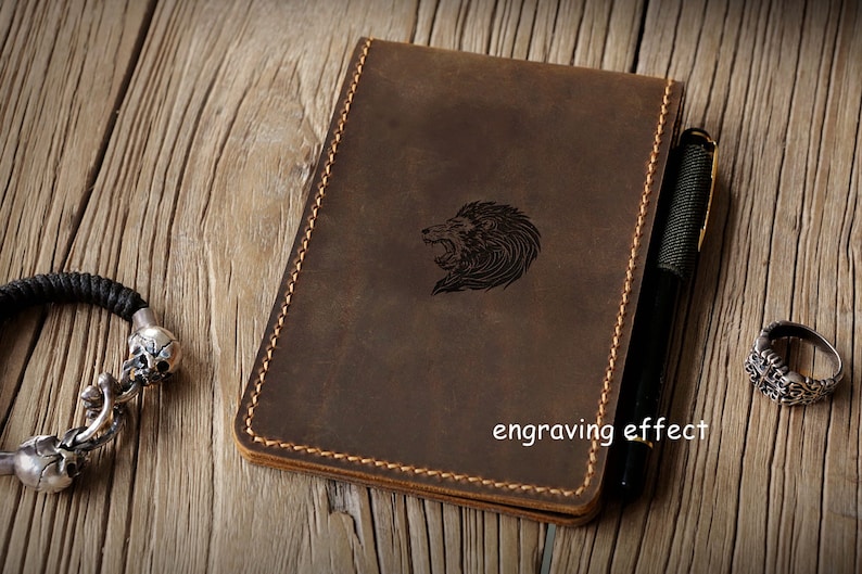 Op de afbeelding: Een bruin lederen notitieboek met een gravure van een leeuwenkop, gestikte randen en een zwarte pen aan de zijkant. Een zwart gevlochten armband met zilveren schedelkralen en een zilveren ring met een kruismotief liggen ernaast.