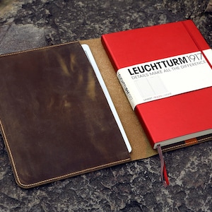 May include: A brown leather notebook cover with a red notebook inside. The notebook has the text "LEUCHTTURM1917 DETAILS MAKE ALL THE DIFFERENCE" on the front.