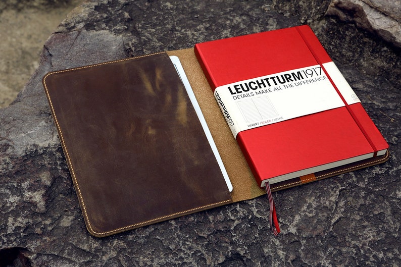 May include: A brown leather notebook cover with a red notebook inside. The notebook has the text "LEUCHTTURM1917 DETAILS MAKE ALL THE DIFFERENCE" on the front.