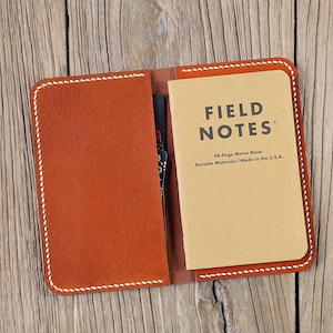 May include: A brown leather notebook cover with white stitching, open to reveal a Field Notes brand notebook with the text "FIELD NOTES 48-Page Memo Book Durable Materials / Made in the U.S.A."