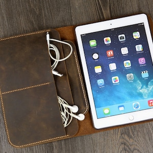 Op de afbeelding: Een bruine leren tas met een klep die opengaat om een witte tablet en een vak voor koptelefoon en snoeren te onthullen.