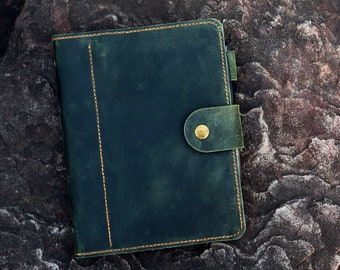 Puede incluir: Una funda de cuaderno de cuero verde oscuro con una línea vertical de costuras naranjas en el lado izquierdo y una correa de cierre de cuero a juego, asegurada con un botón de presión de metal color latón en el borde derecho.