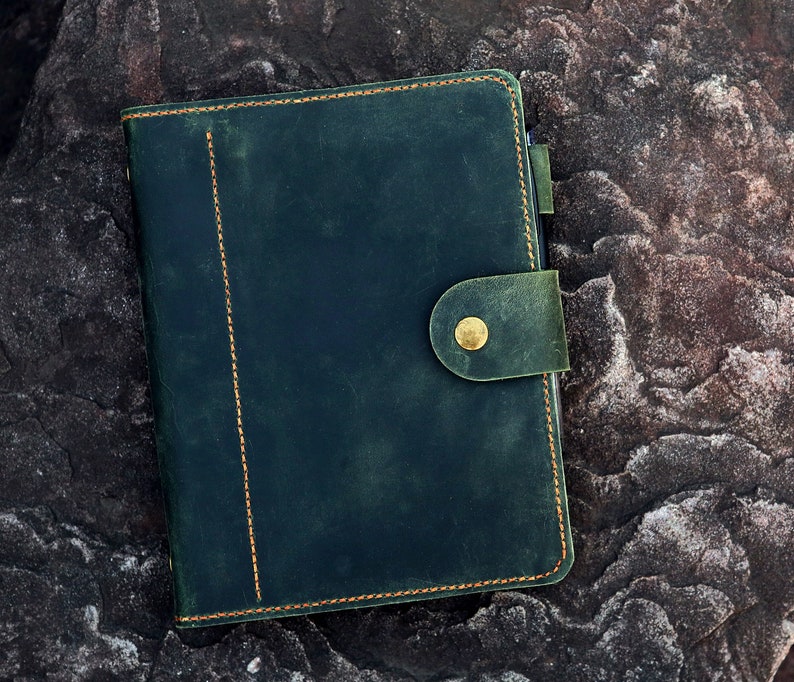 May include: A dark green leather journal with a brass button closure and gold stitching. The journal is lying on a textured rock surface.