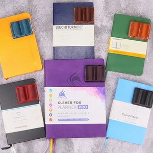 Puede incluir: Seis cuadernos diferentes con portaplumas de cuero adjuntos. Los cuadernos son de varios colores, incluyendo amarillo, azul, verde, morado y negro. Los portaplumas son de cuero marrón, negro y azul.