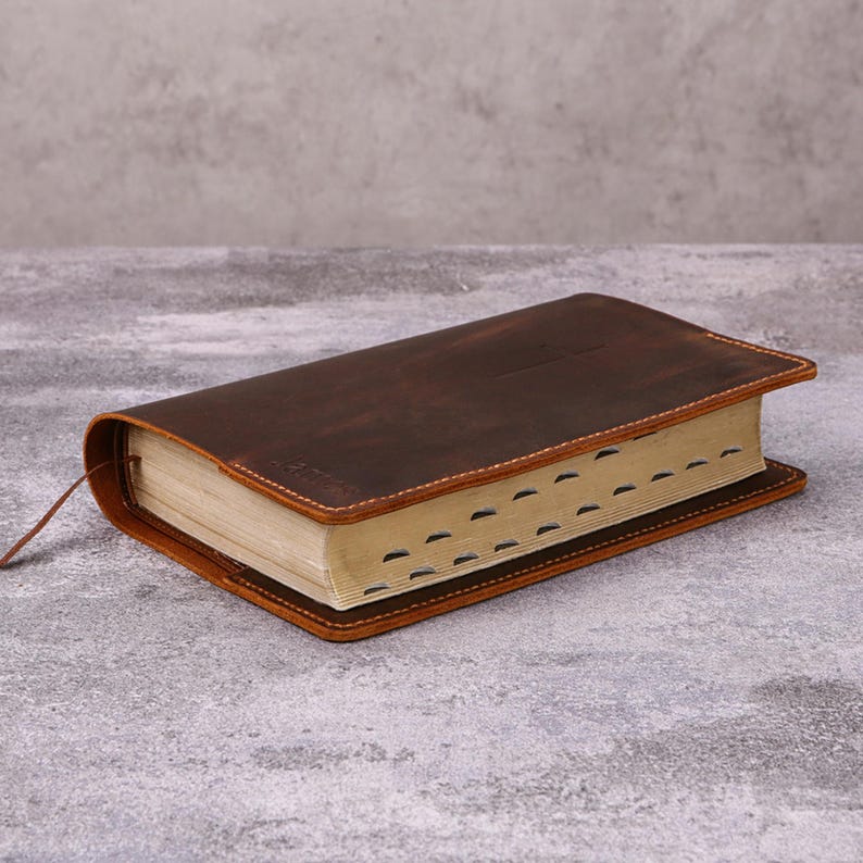 May include: A brown leather-bound book with a textured cover and cream-colored pages. The book has a sewn binding with visible stitching and a brown ribbon bookmark. The edges of the pages are marked with tabs.