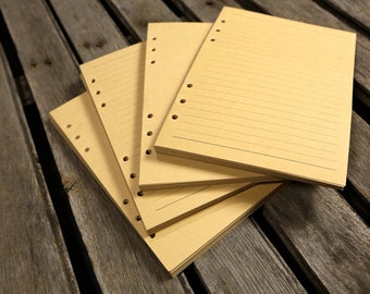 Puede incluir: Una pila de cuatro hojas de recambio de papel marrón con líneas y agujeros perforados para una carpeta. Las hojas están sobre una superficie de madera.