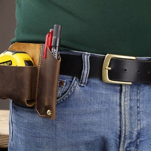 May include: A brown leather tool belt with a yellow tape measure and black and red pens. The belt is worn over blue jeans and a green shirt.