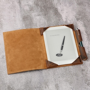May include: A brown leather case with a flap that opens to reveal a white e-reader with the text "For Those Who Write". A green pen is tucked into the case.