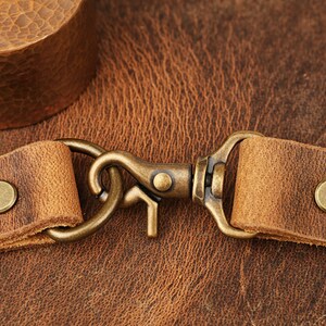 May include: Close-up of a brown leather strap with a brass hook and two brass rivets. The strap is on a brown leather background.