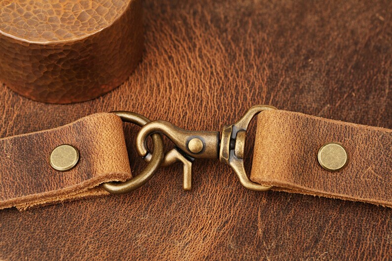 May include: Close-up of a brown leather strap with a brass hook and two brass rivets. The strap is on a brown leather background.