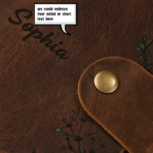May include: A brown leather journal cover featuring a brass snap button closure. The surface displays the name Sophia and a speech bubble containing the text we could engrave your initial or short text here, alongside a small green floral design.