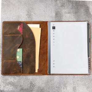 Op de afbeelding: Een bruine lederen folio-hoes met een linker binnenpaneel met meerdere gebogen vakken voor kaarten en papieren, en een rechterpaneel met een digitale tablet met een wit scherm, navigatiepictogrammen en een zwarte styluspen.