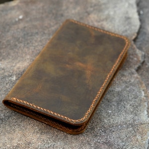May include: A rectangular brown leather wallet featuring a distressed finish and visible tan stitching along the edges. The wallet is folded closed, displaying a smooth, matte surface texture with natural variations in the leather's dark brown color.