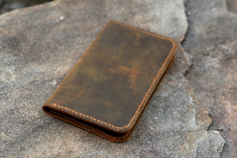 May include: A rectangular brown leather wallet featuring a distressed finish and visible tan stitching along the edges. The wallet is folded closed, displaying a smooth, matte surface texture with natural variations in the leather's dark brown color.